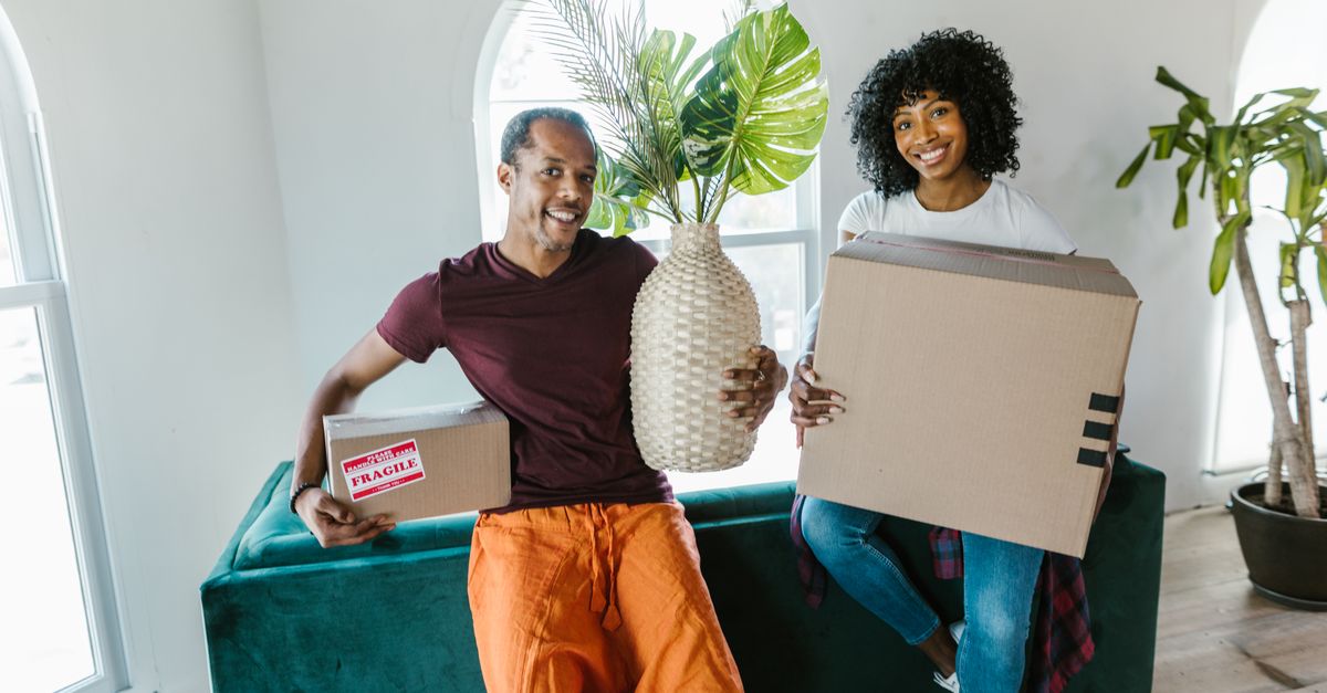 Couple smiling while moving into a new home, carrying boxes and a plant.